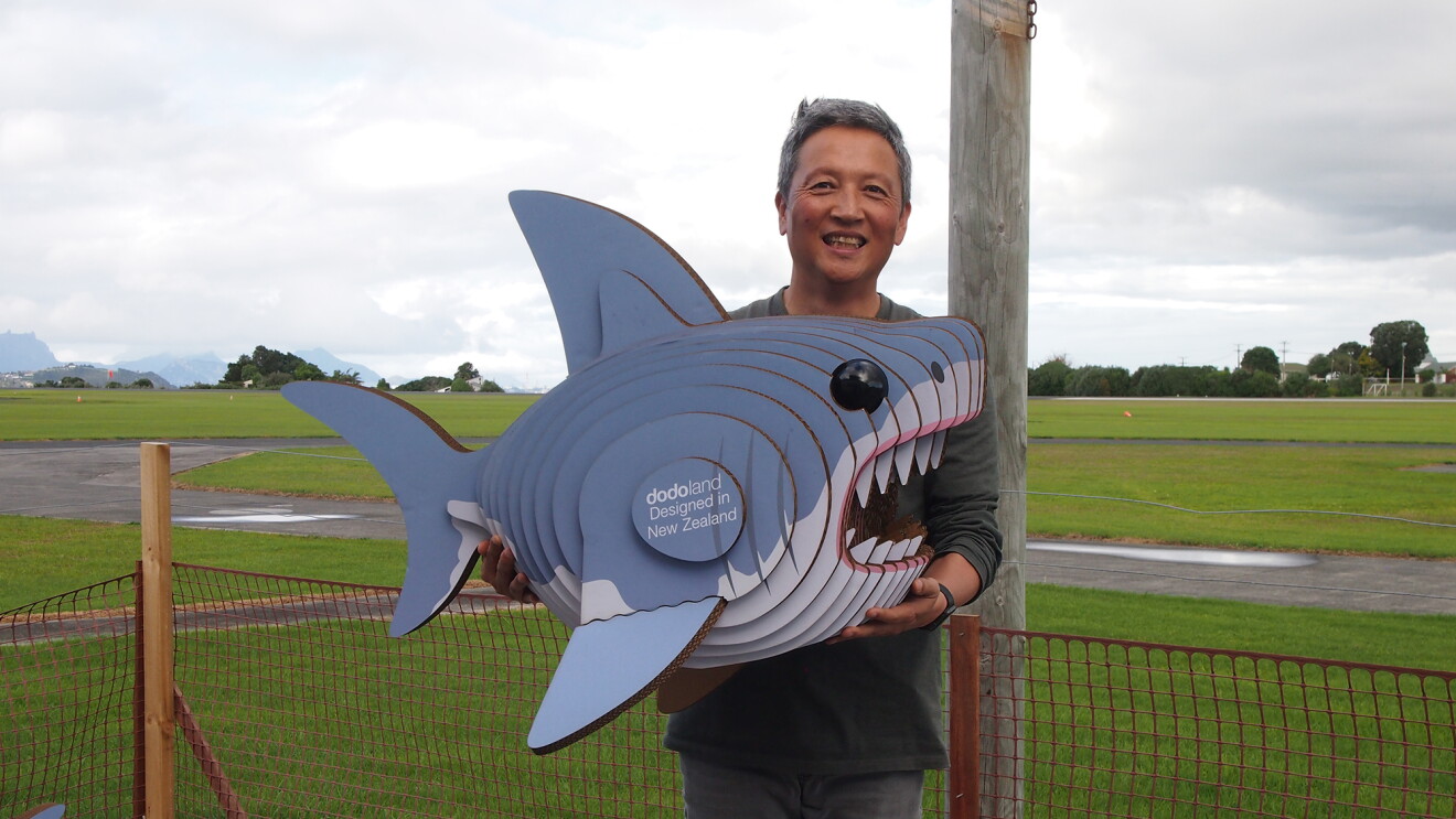 A man stands in a green space, holding a recycled cardboard shark. 