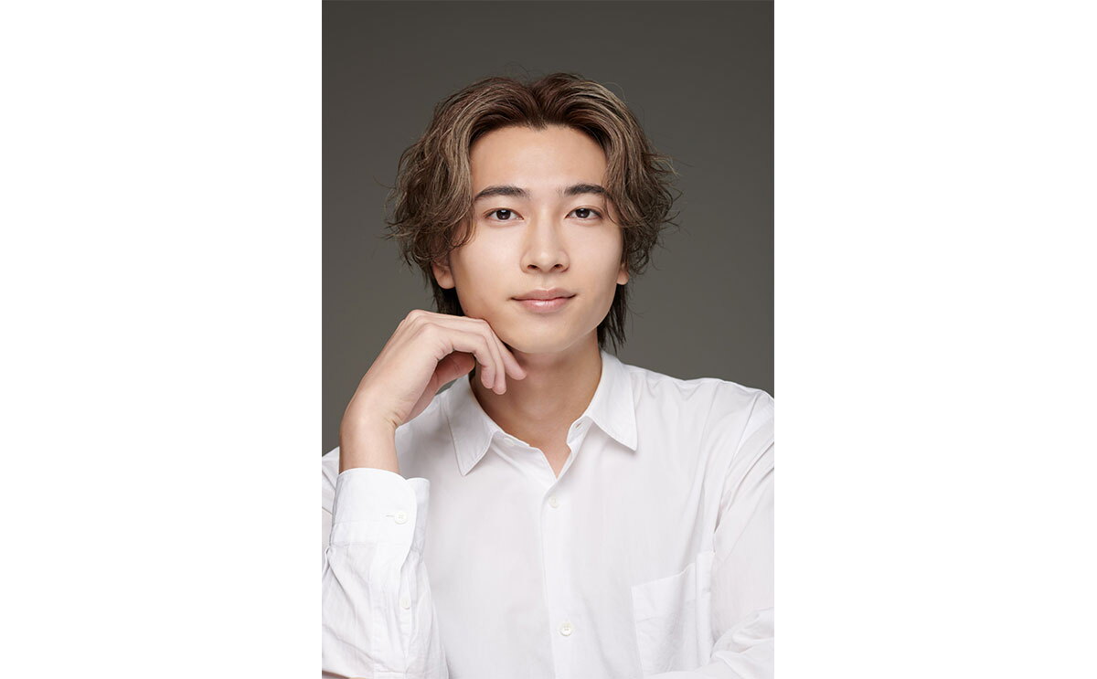 茶髪の男性の上半身ポートレート。白いシャツを着用、右手を軽く顎に添えている。