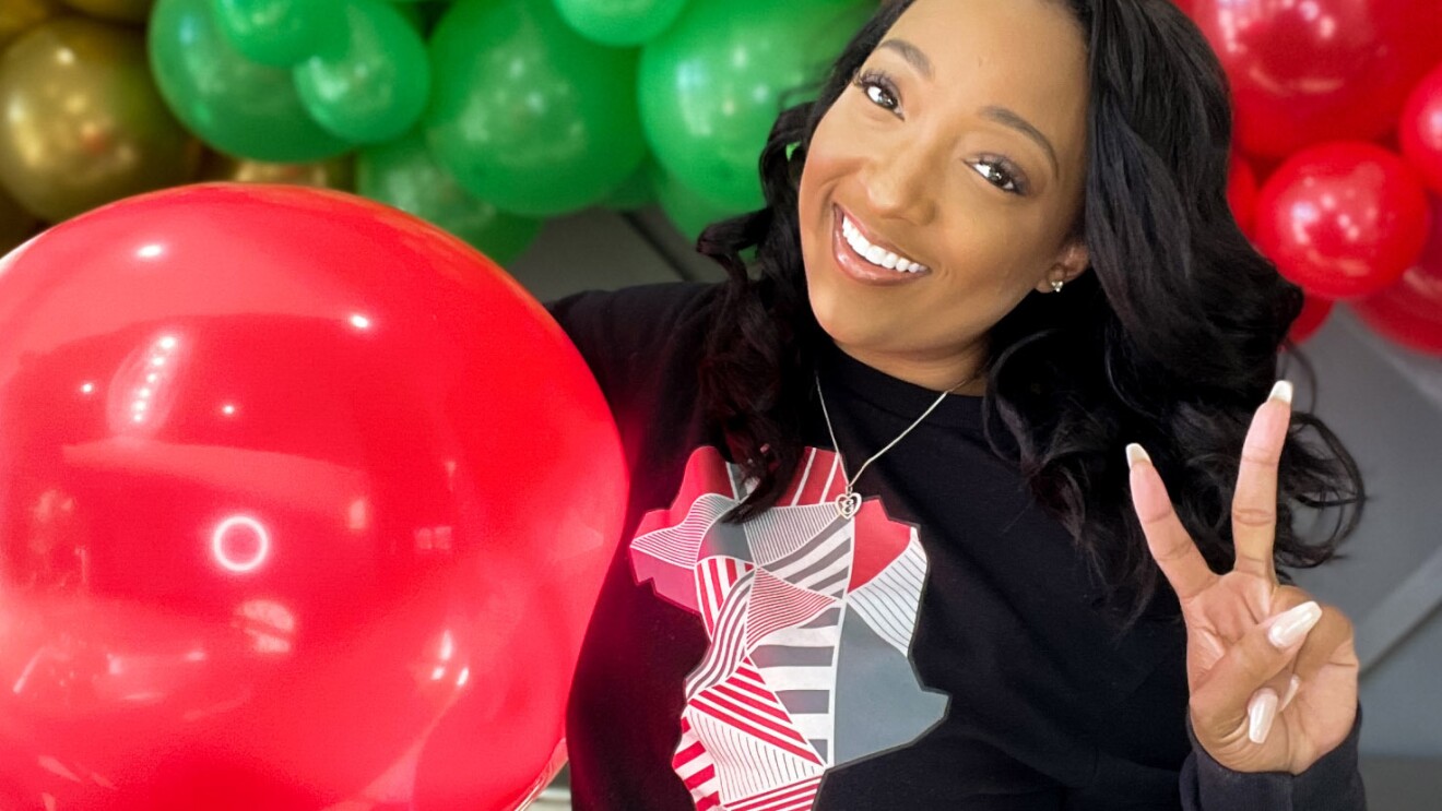 An image of a woman smiling for a photo while making a peace sign. She is standing in front of a balloon arch with green, red, black, and gold balloons, and holding a large red balloon under one arm.