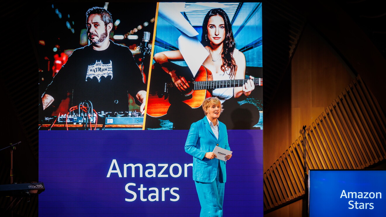 En un escenario aparece Tania Llasera vestida con un traje chaqueta azul eléctrico y una camisa azul con un papel en la mano. De fondo hay un televisión con un fondo azul y el texto Amazon Stars. Y muy detrás, en el escenario hay un pantalla de 3 x 3 metros con la parte inferior de azul y el texto Amazon Stars y la parte superior, partida en dos, con la foto de Obed Soto y Marta Mallo. Obed con el pelo corto y barba está delante de una tabla de mezclas de sonido y Marta está sentada en el suelo tocando la guitarra. Él va vestido con una camiseta negra con la figura de Batman en blanco y ella con un pantalón negro y una camiseta blanca.