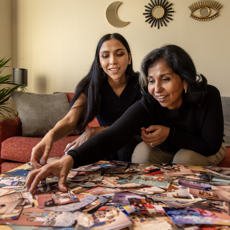 Two women sitting on a couch reach toward a table full of old photos.