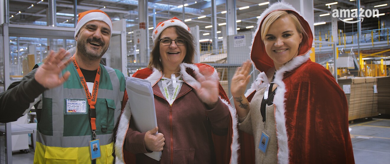 3 Mitarbeiter winken in die Kamera, alle tragen eine rote Zipfelmütze. Der rechte Mitarbeiter trägt ein Band mit Aufschrift "Betriebsrat".