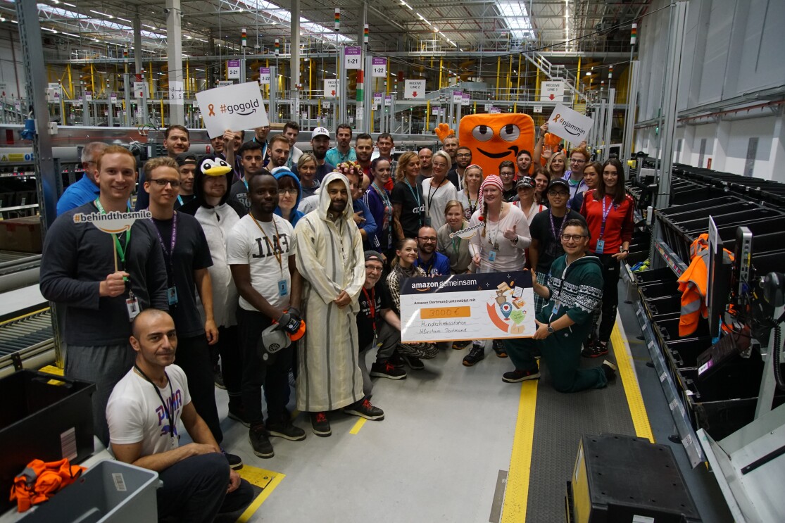 Rund 30 Mitarbeiter in Schlafanzügen haben sich in einer Logistikhalle fürs Gruppenbild versammelt. Alle gruppieren sich um einen großen Spendenscheck, der von einigen Mitarbeiter gehalten wird.