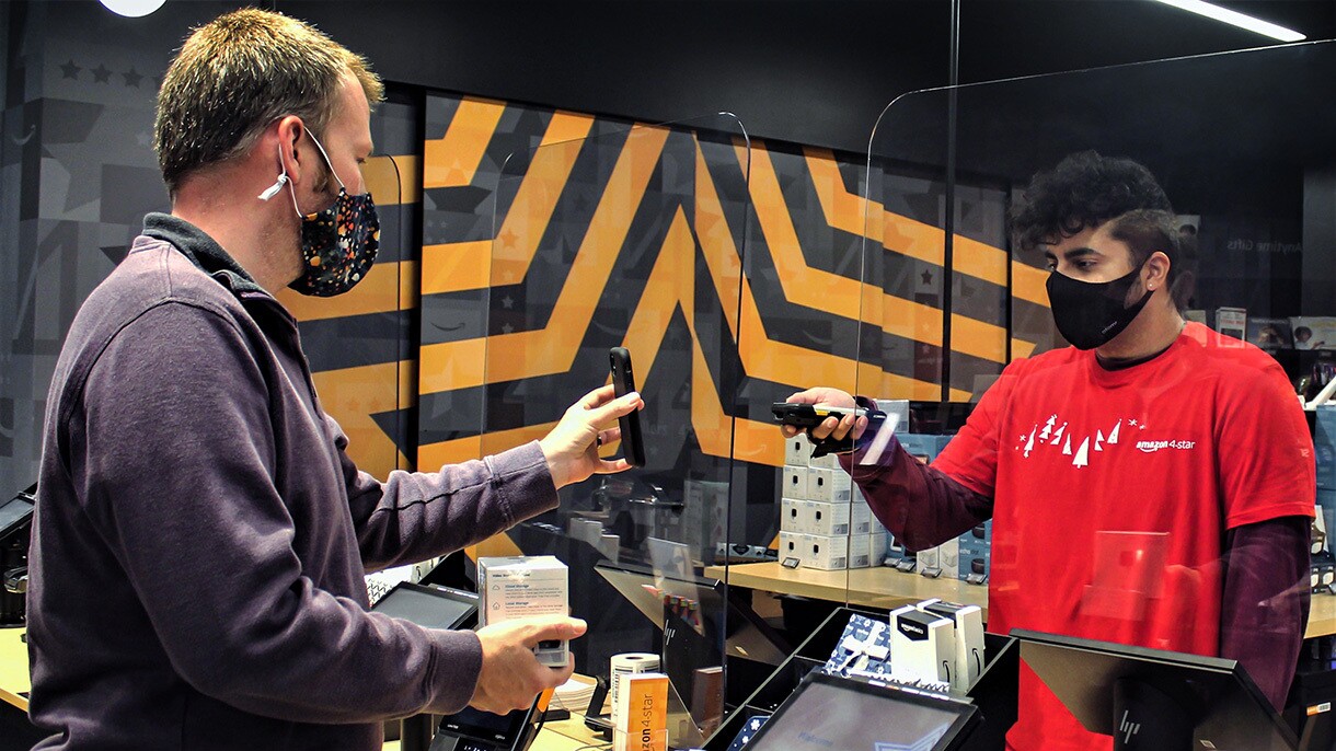 A man stands at an Amazon store counter, holding up his phone so a store associate can scan the barcode to facilitate a return.