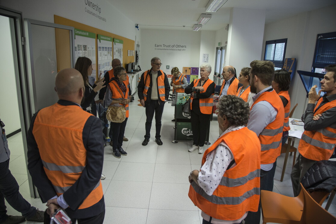 Gruppo di nonni di dipendenti Amazon in visita al Centro di distribuzione urbana Prime Now di Milano in occasione della Festa dei nonni. Indossanno giubbini catarinfrangenti arancioni. Sui muri della sala in cui si trovano si leggono due principi di leadership di Amazon: 'Earn trust of others' e 'Ownership'