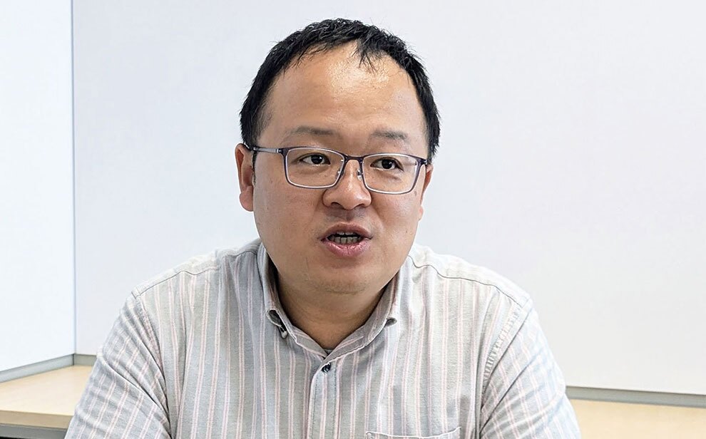 白い壁を背景に、眼鏡をかけて縦じまのシャツを着用した男性が話している。