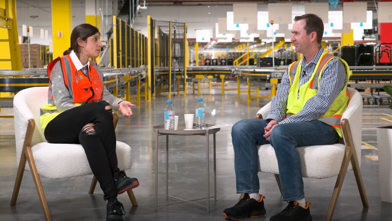 John Felton, vicepresidente Senior de Operaciones Mundiales de Amazon, entrevista a la directora general del mayor centro logístico de Madrid. Los dos están sentados en unos sillones blanco dentro de un centro logístico. 