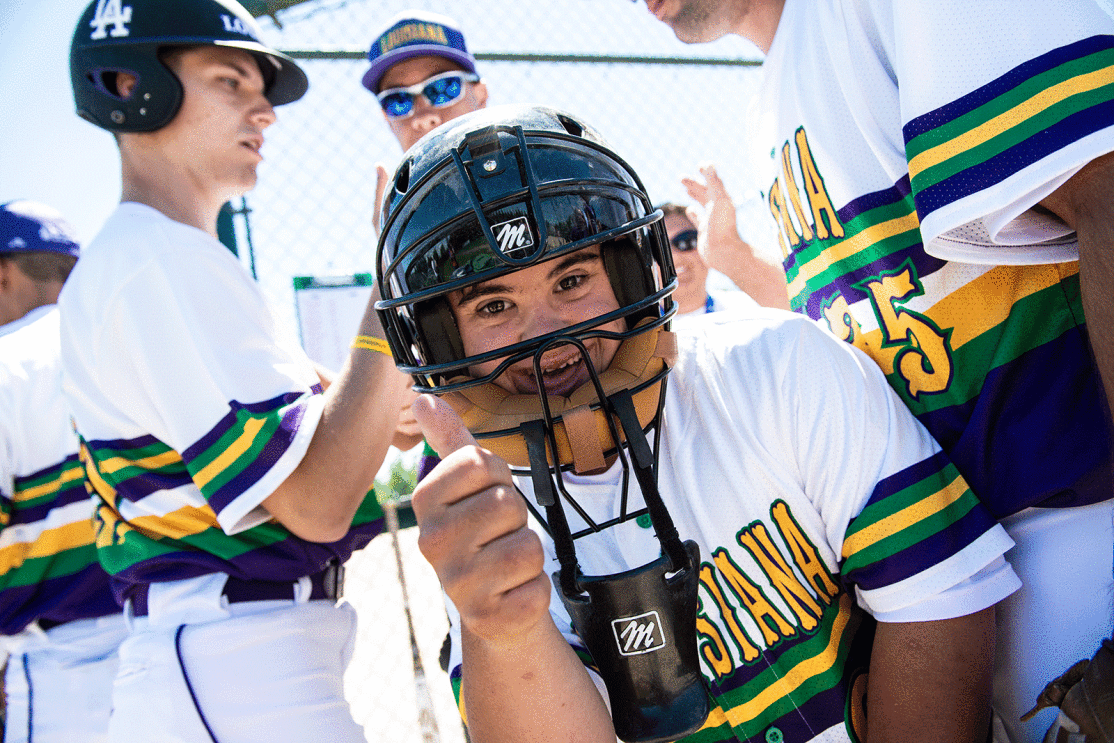 An image of a person wearing a baseball helmet and uniform giving a thumbs up. The background shows other people in the same uniform doing a handshake. 