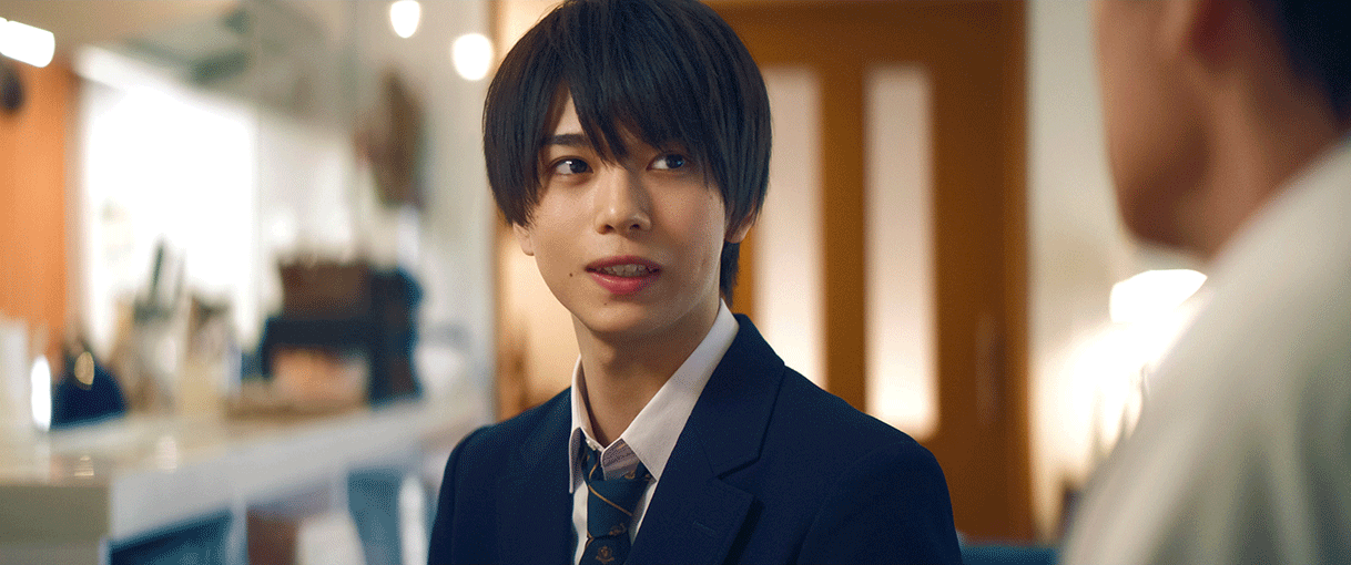 奥にキッチンとドアが見える室内で、右方向を見る制服の少年。少年の視線の先にもう1人人物がいるが、ピントがぼやけている。