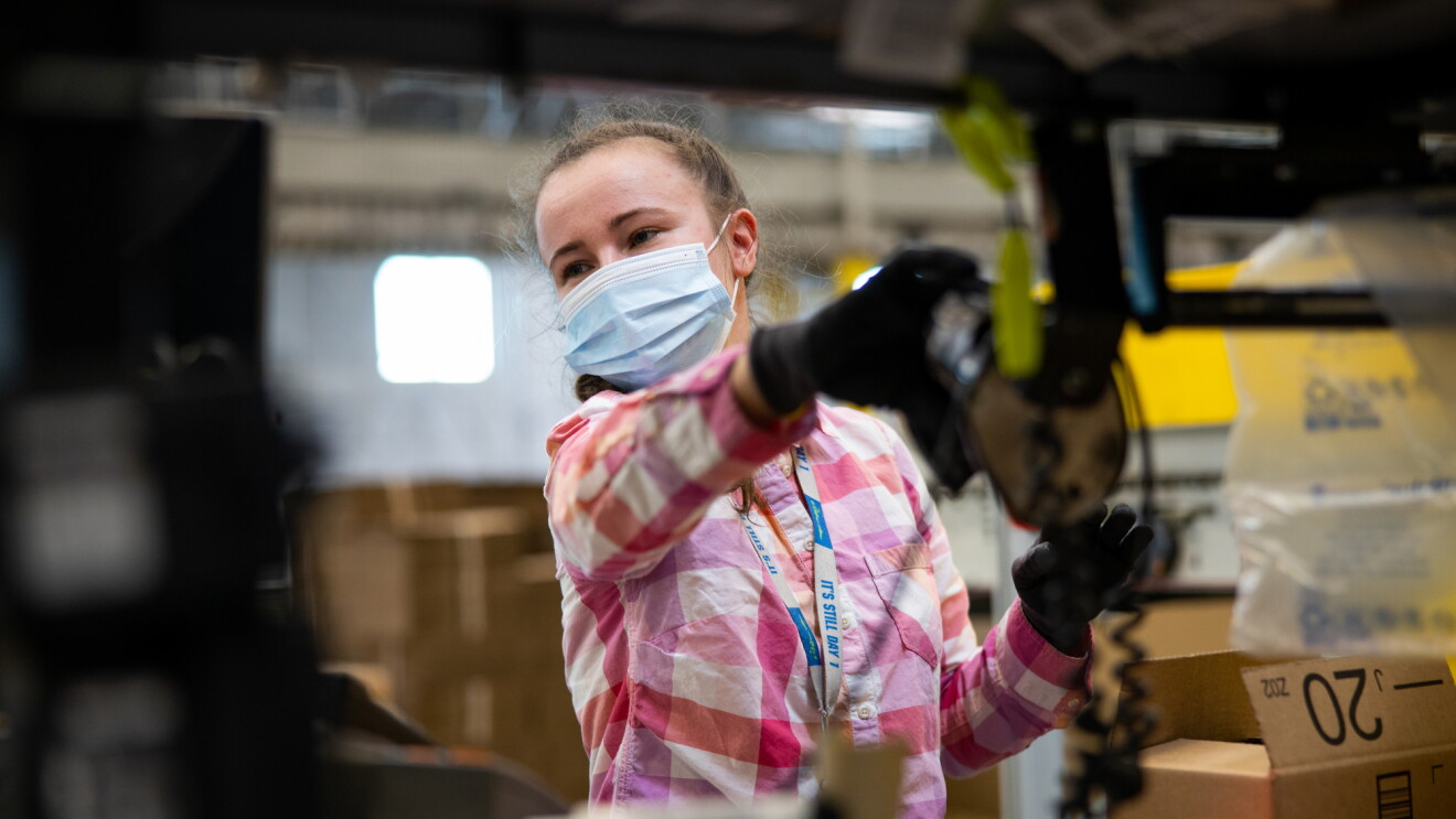 A woman works while wearing a mask. To her left, there is a cardboard box with the Amazon logo.
