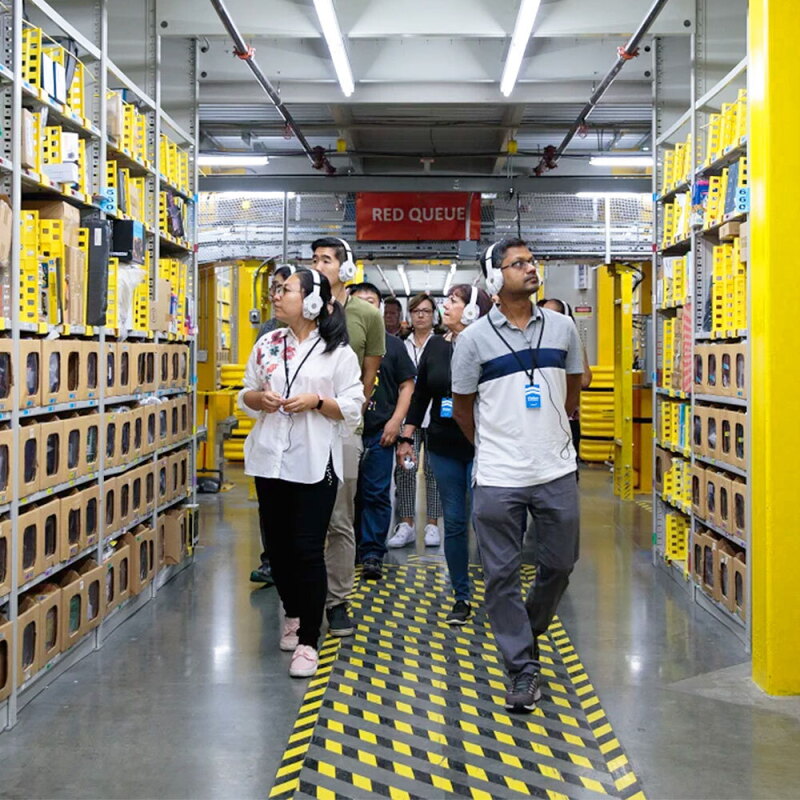 Group of people wearing headphones and walking down an aisle during an Amazon fulfillment center tour.