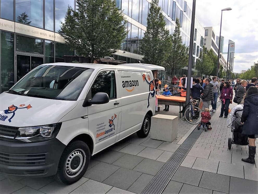 Ein weißer Lieferwagen mit Aufdruck "SystemLogistik" und "Amazon" parkt vor einem Bürogebäude.
