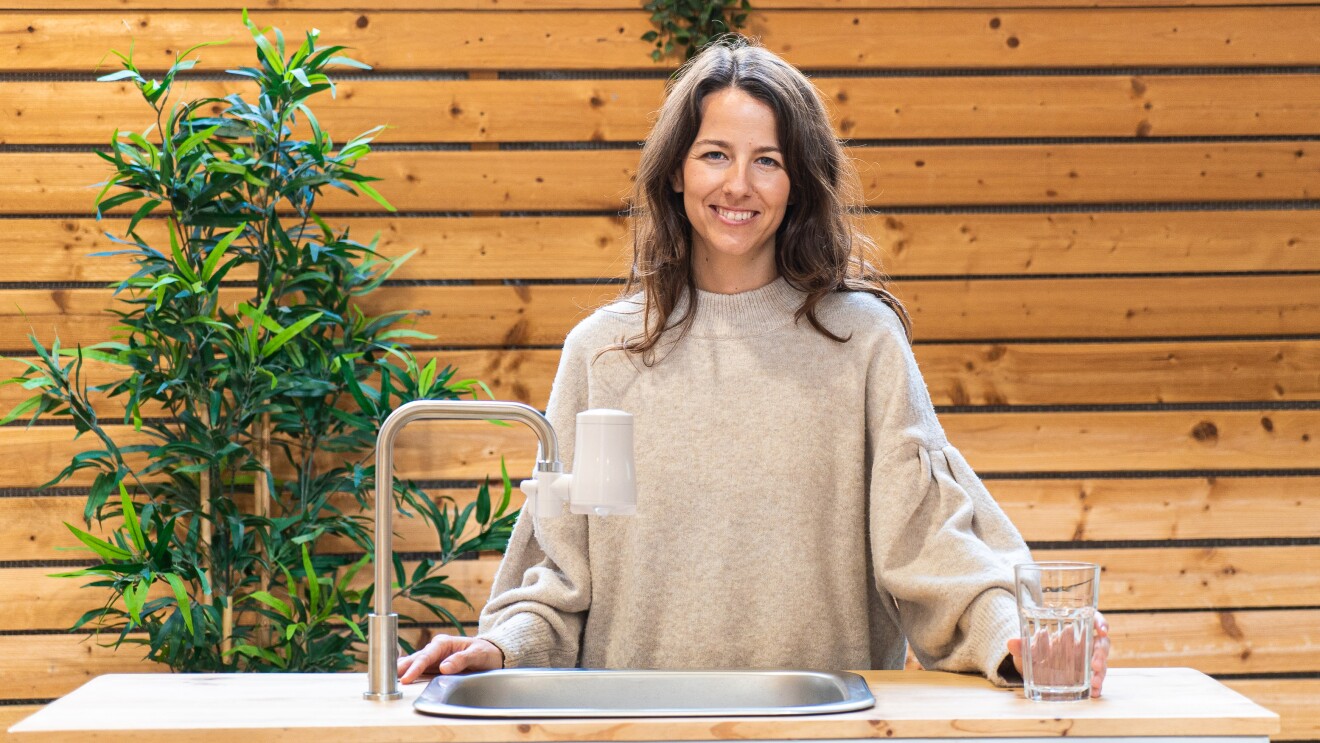 La start-up TAPP Water fue fundada por Magnus Jern y Alexander Schwarz. En la foto aparece la CEO Rocío Alcocer en una mesa con un gripo que tiene el filtro de Tapp water . Rocío va un jersey también marrón muy claro. Tiene el pelo largo castaño y las manos apoyadas sobre la mesa, sujerta una vaso de cristal con agua. De fondo una pared de madera.