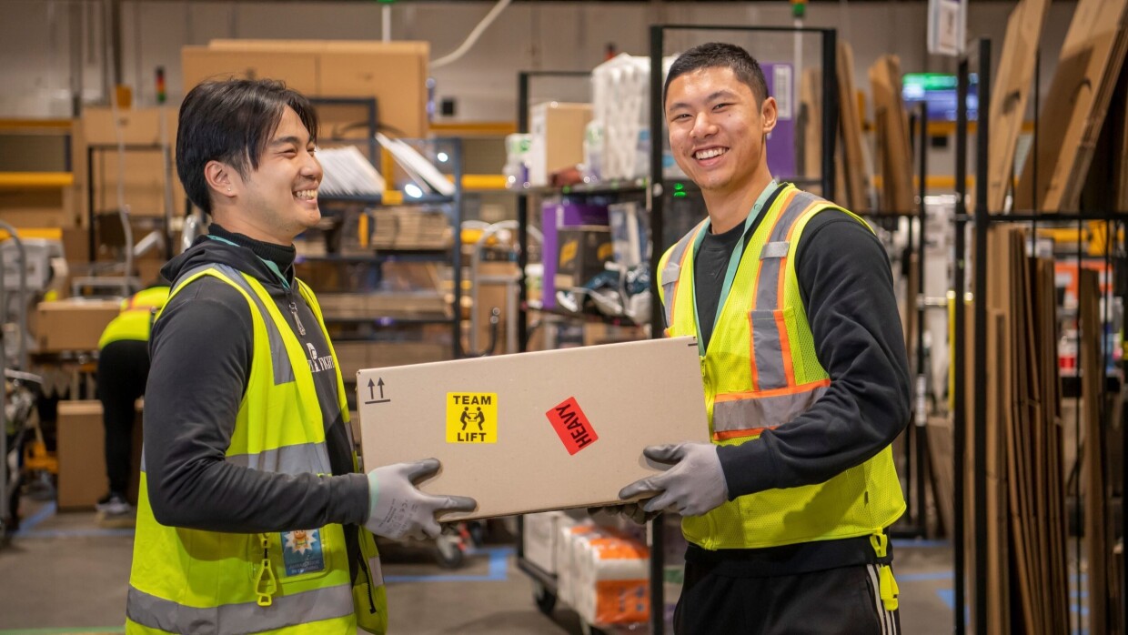 two men wearing safety vest are lifting a box together in a warehouse