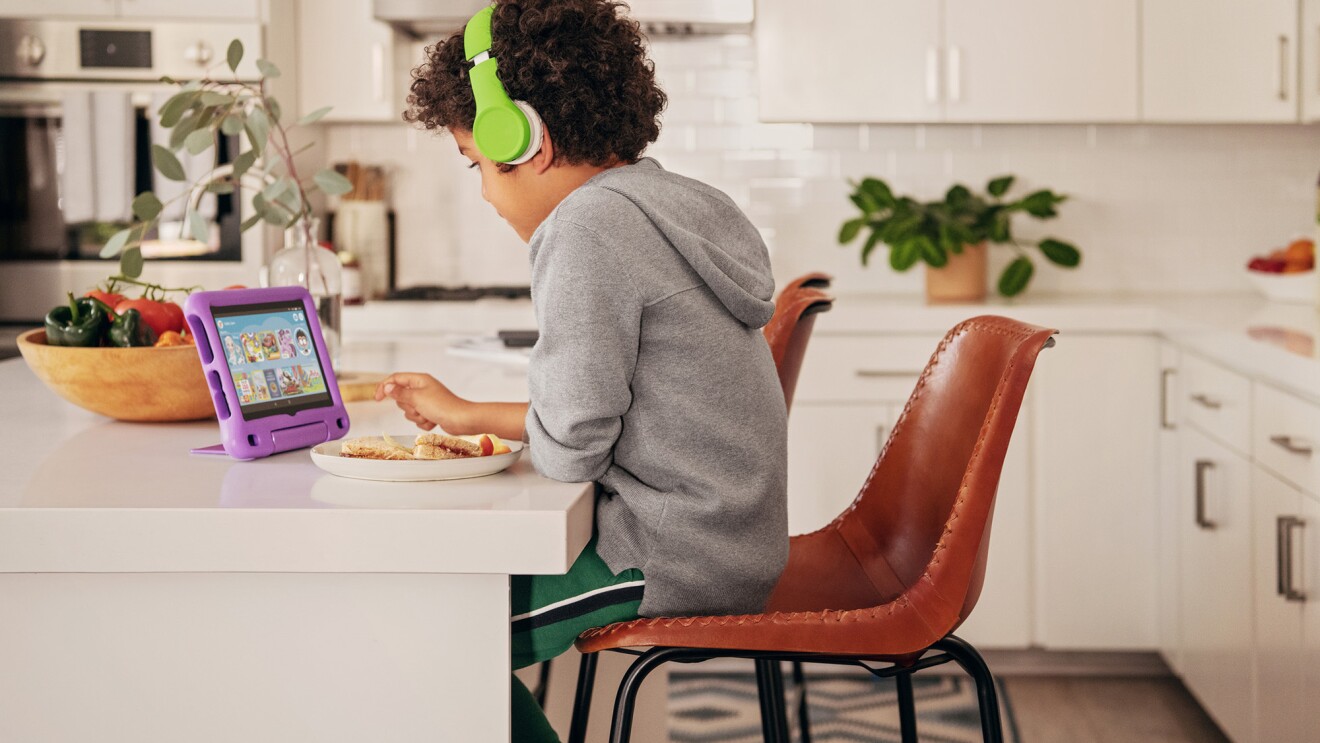 A child sits at a kitchen island, looking at a fire tablet. He wears headphones as he uses the device.
