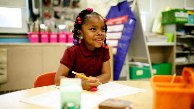 A young girl in a school setting sits with a pen and paper. Blurred out in front of her is a milk carton and snack.