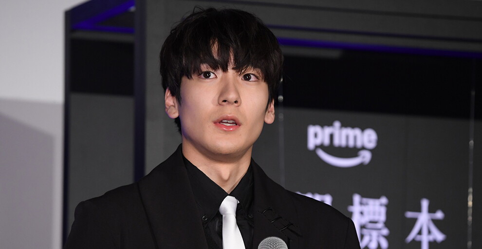 黒と白の壁を背に立つ青年。黒いジャケットに黒いシャツ、白いネクタイを着用。