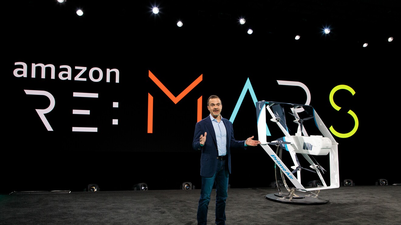 Jeff Wilke, CEO consumer worldwide at Amazon, stands on a stage. Behind him is a re:MARS logo. To his left is a drone painted in white and light blue.