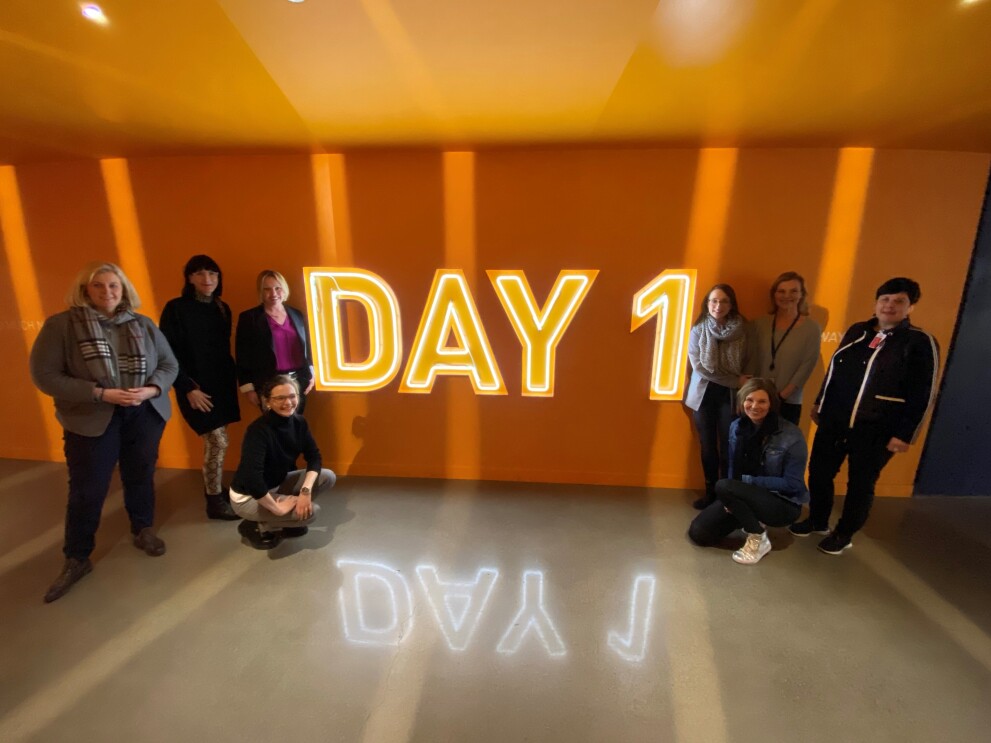 Gruppenfoto vor einer orangefarbenen Wand mit dem leuchtenden Schriftzug "Day 1".