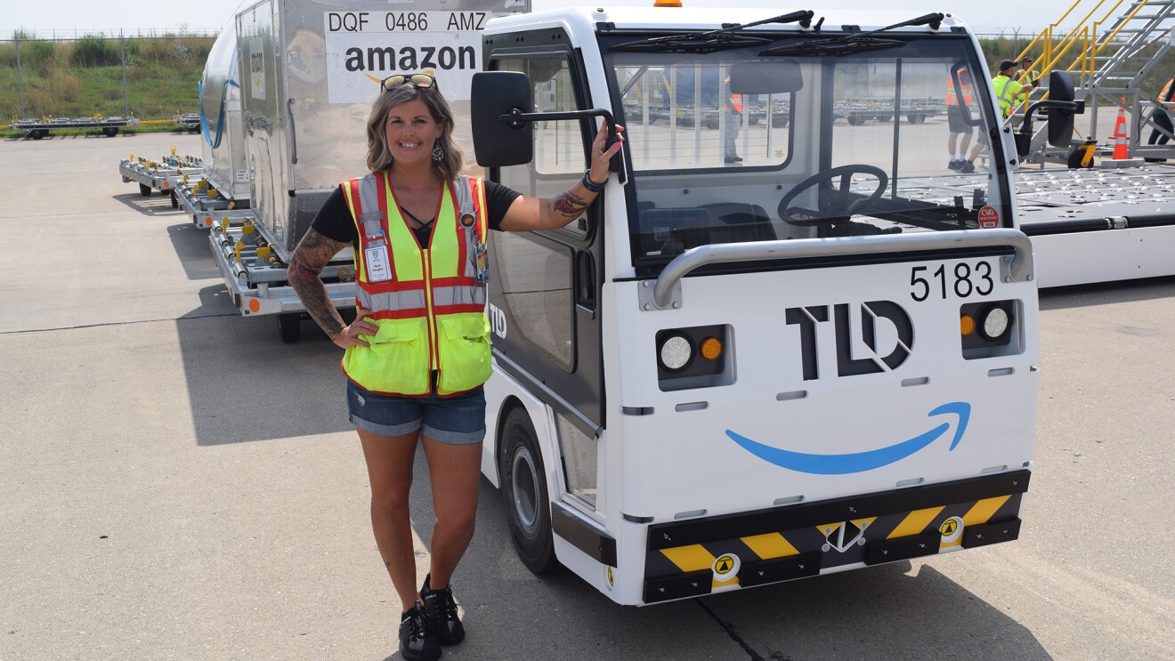An image of a woman standing in front of a truck that is pulling large metal containers behind it. She is smiling for the photo and wearing a safety vest while working at the Amazon Air Hub in Kentucky.