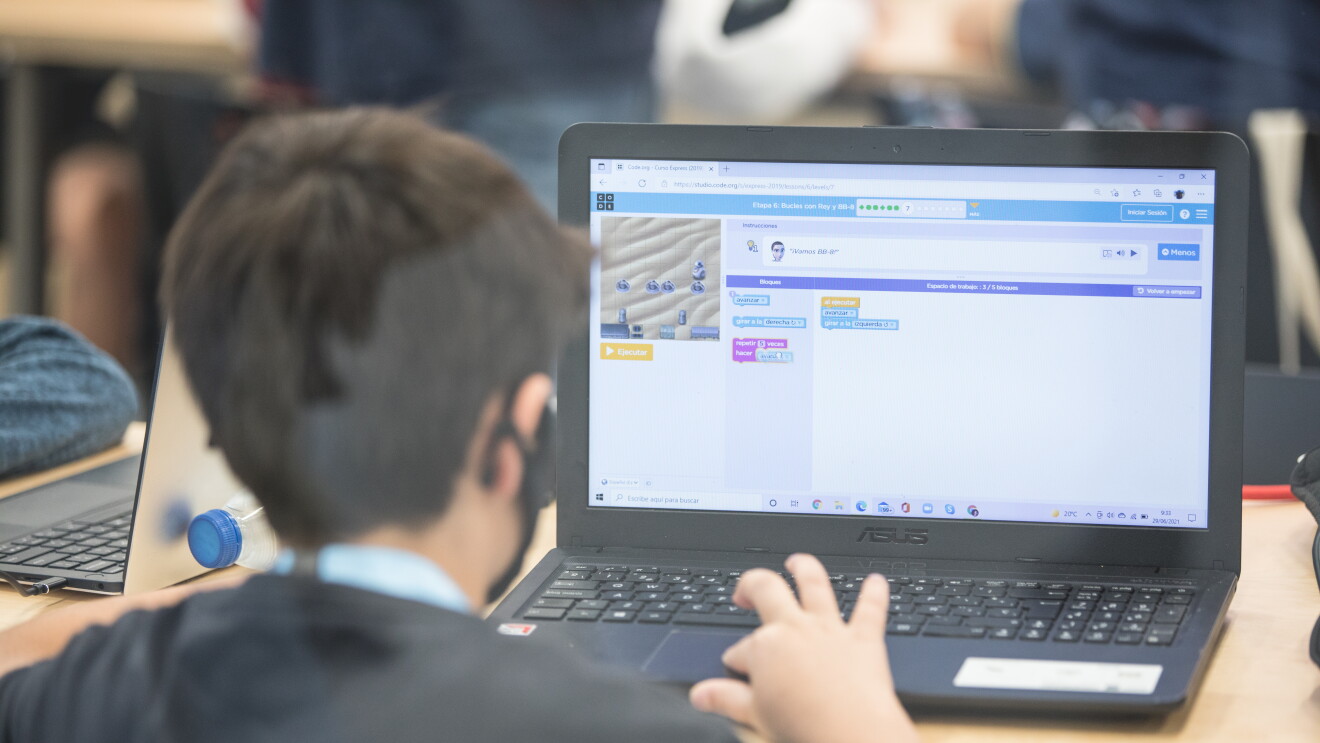 En una mesa de un centro educativo, aparece un niño de espaldas, con una camiseta negra, una mascarilla y el pelo corto marrón con la mano derecha apoyada en el teclado de un ordenador portátil. La pantalla del ordenador está desenfocada. En la mesa del chico aparece una botella de plástico tumbada y otro ordenador portátil de un compañero.