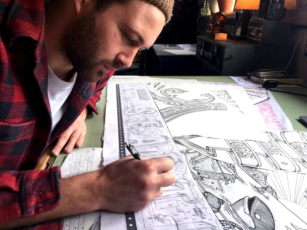 Kyler Martz in his Seattle studio, drawing the word Seattle onto a piece of sketch paper. He is wearing a red and black checked flannel shirt. 