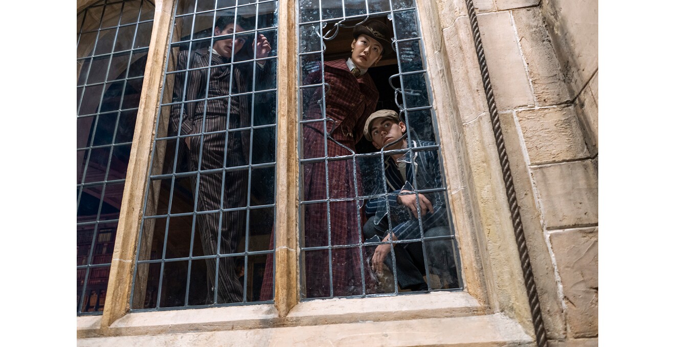 ベージュ色の石壁の大きな窓を屋外、下から見上げたところ。窓ガラスの右端が大きく割れていて、金属の格子もねじ曲がっている。内側から3人の人物が窓を調べている。