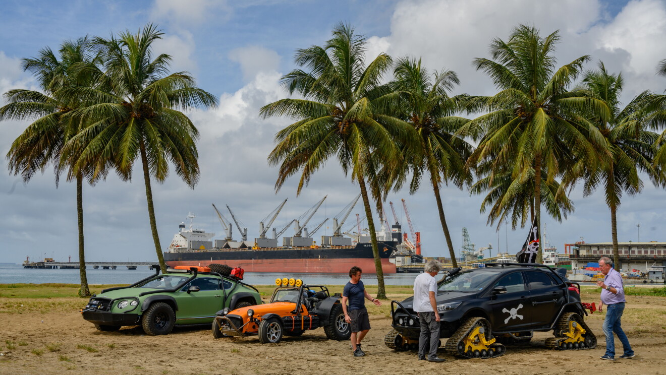 Three men stand next to three cars on a beach with palm trees in the background