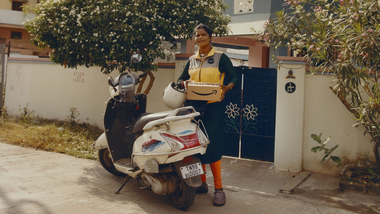 Repartidora de India. Está delante de una casa con la puerta azul, de pie, agarrando una caja de Amazon y con el casco en la mano izquierda. Va con un chaleco Amarillo de Amazon, una falta azul y unos pantalones debajo de color naranja. Delante tiene una moto de color blanco.