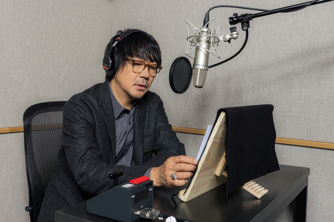 収録ブースで、ヘッドフォンを着けてマイクに向かう、グレージャケットの男性。机の上に書見台がある。手元に赤いスイッチ付きの機器。