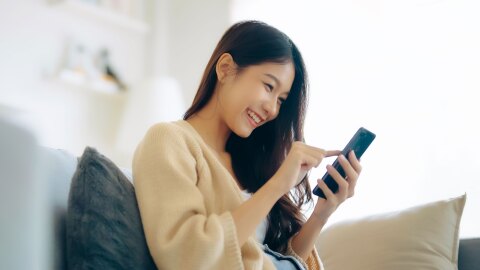明るい光が入る白い背景、クッションにもたれて座る人物。長い黒髪でベージュのカーディガン着用、スマートホンを笑顔で操作している。