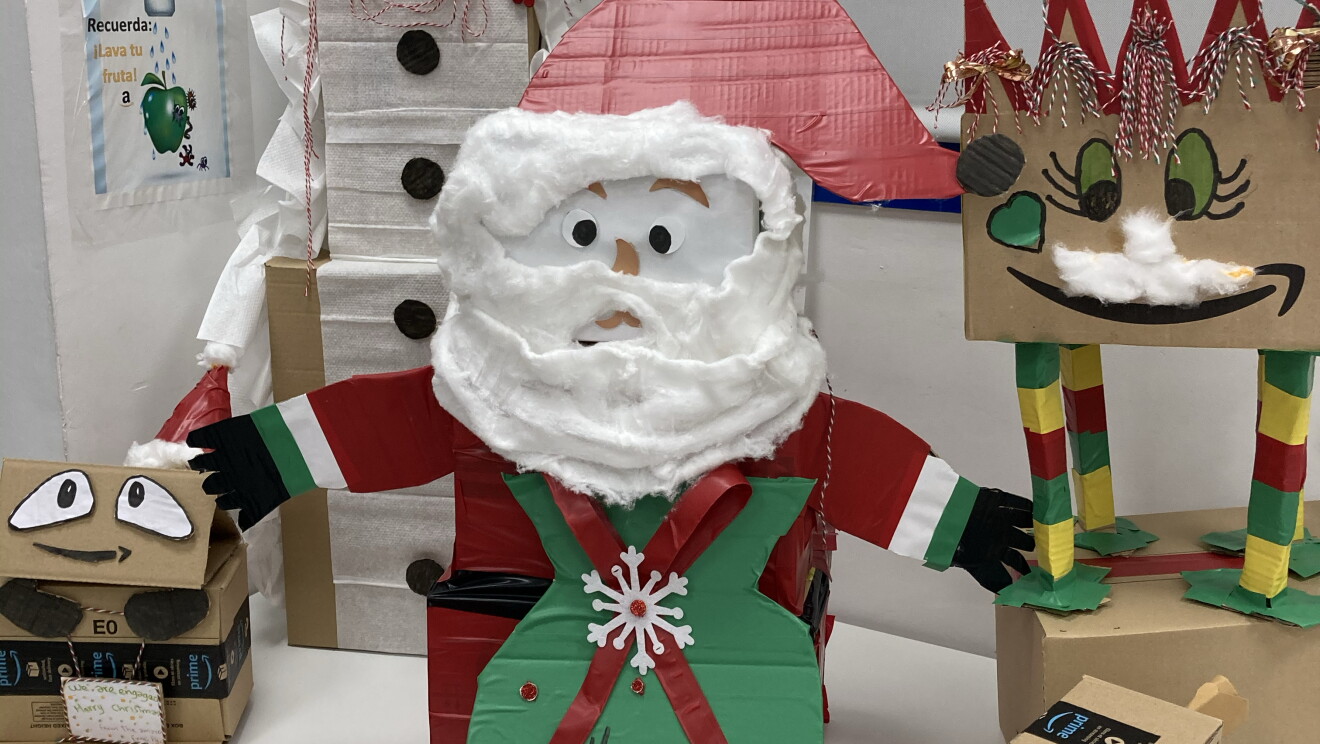 Adorno navideño de DMA1. Encima de una mesa de color blanco hay diferentes decoraciones navideñas hechas con cajas de cartón: un papá Noel, un elfo y un muñeco de nueve. 