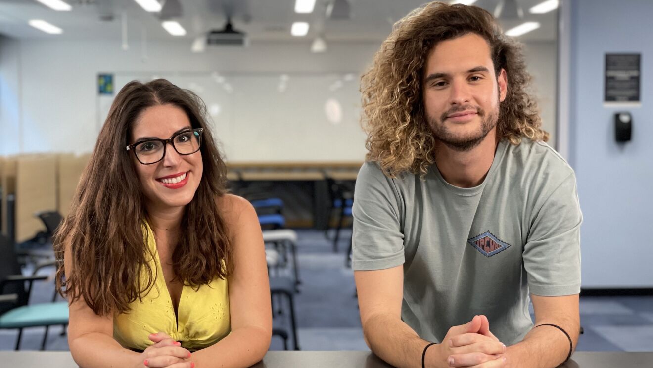 Maria Viñambres y Andrés de Cossío mirando a cámara con los manos agarradas y el antebrazo apoyado en una mesa de color gris. María tiene el pelo castaño ondulado y suelto, las uñas rosas, unas gafas de pasta y un vestido de color amarillo. Andrés tiene el pelo largo rizado por el hombro y suelto. Camiseta de algodón de manga corta y de color gris. En cada muñeca lleva una goma de pelo. Tiene también barba y perilla de dos días. De fondo se ve una sala con sillas y mesas de tonos azules y verdes y una pizarra blanca de fondo.