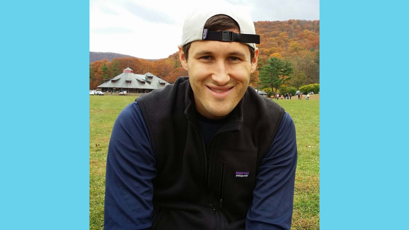 Harrison smiling while sitting in a grassy field with colorful fall trees in the background.