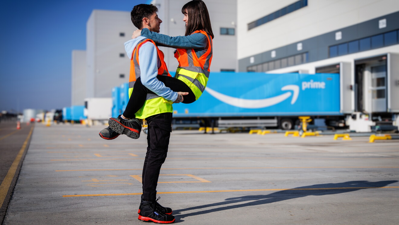 Manel&Laura, Amazon Illescas.Él le agarra y de fondo se ve un camión de Prime y el centro logístico de Illescas.  