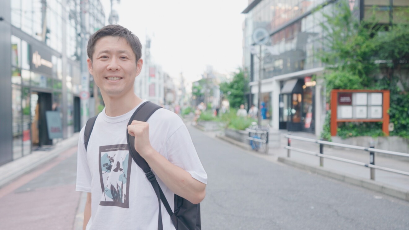 白いTシャツを着て黒いバックパックを持っている男性（可部直人さん）がショッピングエリアの近くに立っている。