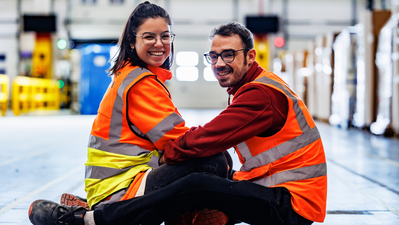Marta y Roberto posando en MAD6 para la sesión de fotos del post que tenemos entre manos. Los sentado uno frente al otro, él con las piernas con fuera y ella por dentro, . Miran a cámara. Ella lleva media melena castaña con una coleta alta y el pelo medio suelto. Los dos llevan gafas y él barba y bigote de dos días. Ella una chaqueta de color naranja de seguridad y un chaleco y él un chaleco con un polar de color rojo debajo. De fondo desenfocado un pasillo de un centro logístico. 