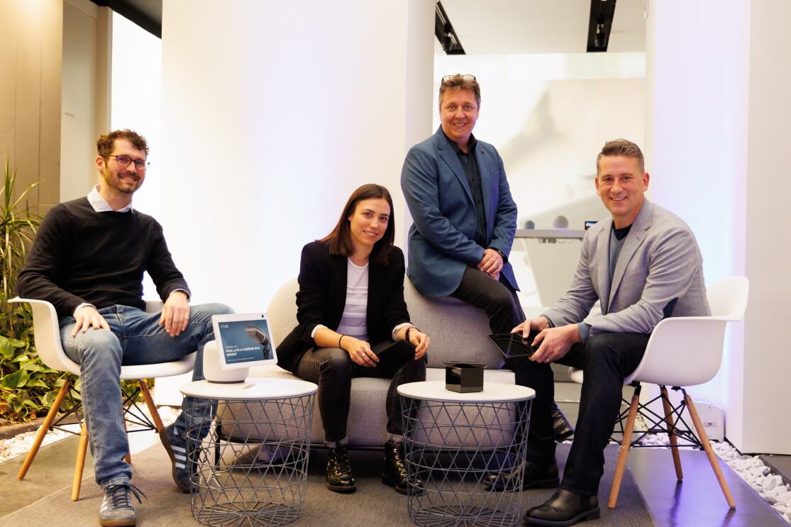 Equipo de Amazon Lab 126 en España. En la foto, el interior de un espacio de descanso de unas oficinas. Aparecen cuatro personas; tres hombres y una mujer. Están todos sentados. En prímera línea hay dos mesas donde en una hay un dispositivo de Amazon. En el centro hay un sofá de dos plazas con la mujer sentada y un hombre apoyado en el respaldo. En el límite izquierdo y derecho del sofá hay dos sillas blancas con las otras dos personas sentadas.   Descripción de todos las personas que aparecen de izquierda a derecha. El primero lleva unas deportivas, unos jeans, una camisa con un jersey, gafas y el pelo corto y barba de dos días. La mujer va con unas botas, un patalón y una americana negra. Debajo lleva una camiseta blanca. El pelo largo marrón. El segundo hombre lleva pantalones negros y americana y camisa azul oscura. Tiene perilla blanca, pelo entre rubio y blanco y las gafas en la cabeza. El tercer hombre lleva botas y pantalón negro, una americana de color gris y un jersey de punto debajo de color azul. El pelo corto y rubio. En la mano él y la mujer están sujetando un dispositivo Amazon. 
