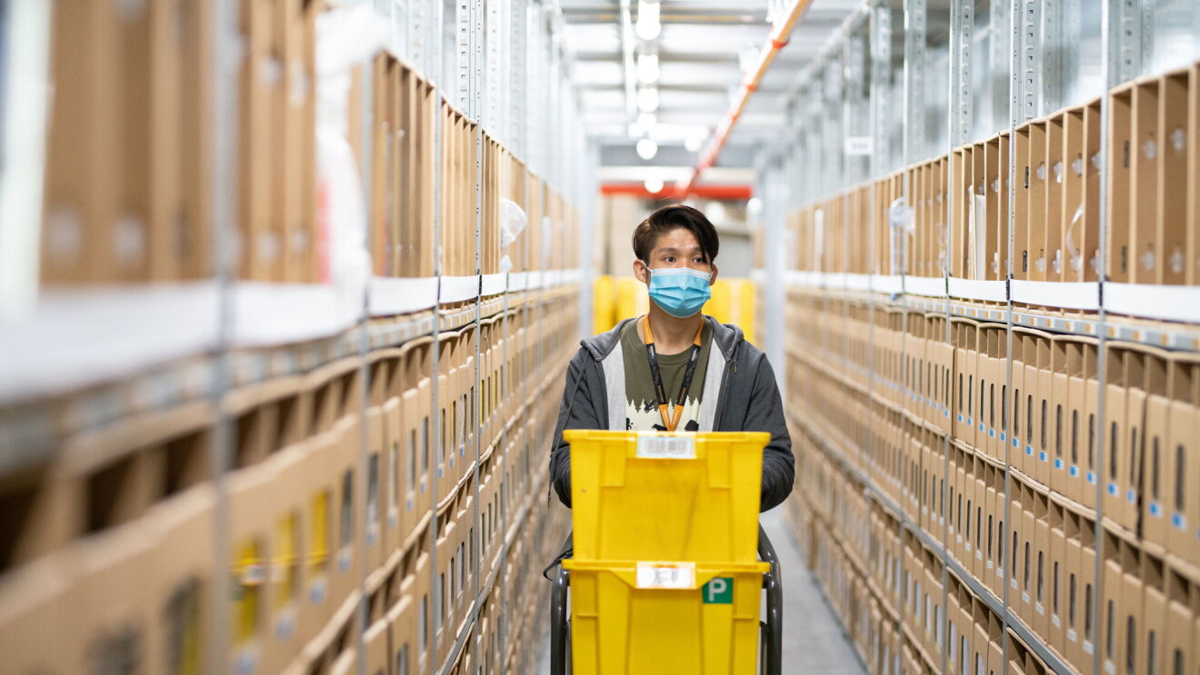 An Amazon employee wearing a face mask pushes a cart with yellow boxes through a fulfilment centre aisle.