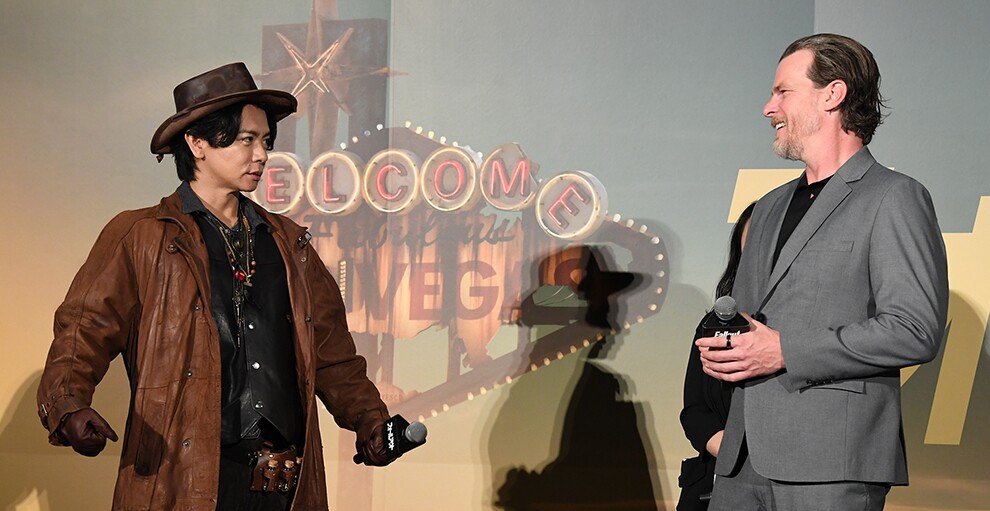 朽ちたネオンが描かれた壁を背に立つ2人の男性。左側の茶色い帽子とコートの男性は両腕を体の横で構えたポーズ。グレーのスーツの男性はそれを見て微笑む。