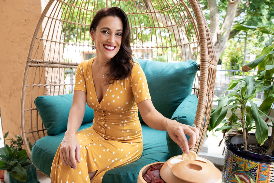 An image of a woman sitting in a wicker chair outside while smiling for a photo in a yello dress with white polka dots. The decor around her is colorful.