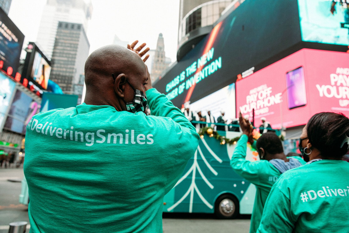 An image of a man clapping and smiling while wearing a mask and watching a choir perform on top of a double-decker bus.