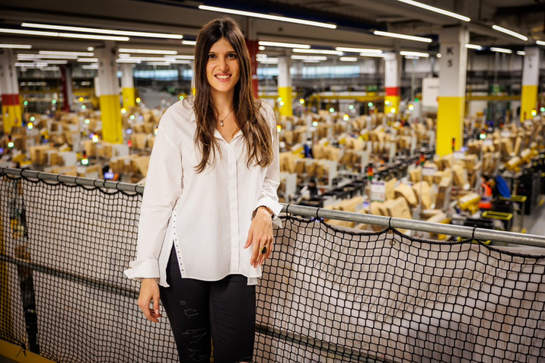 Geraldine Leiros, directora general del centro logístico robotizado que Amazon tiene en Illescas (Toledo). Dentro de un centro logístico aparece del fondo desenfocado con estantería de paquete. Geraldine aparece en un balcón con una red que protege la barandilla de color negro. Ella se encuentra de pie, con una camisa blanca por el muslo y unos pantalones negros. Lleva el pelo largo castaño a la altura del pecho, con un anillo negro grande en la mano derecha y un collar. 