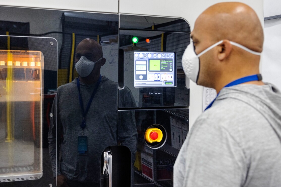 Mahale wears a face mask and stands in front of a 3D printer.