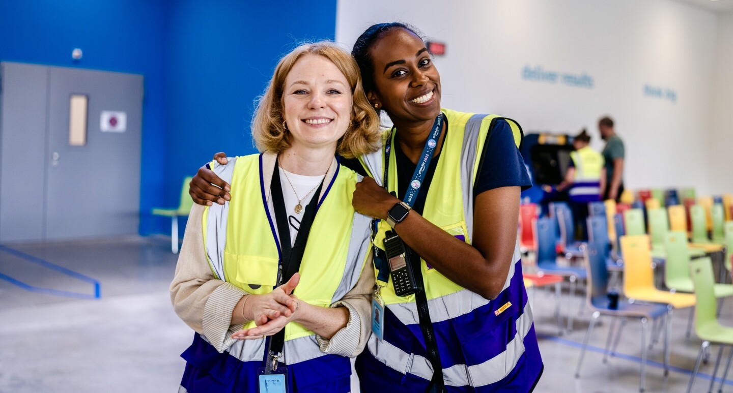 Zwei junge Amazon Kolleginnen aus Kaiserslautern mit Sicherheitswesten halten sich lächelnd im Arm: Die eine ist blond und hellhäutig, die andere ist dunkelhäutig mit dunklen Haaren.