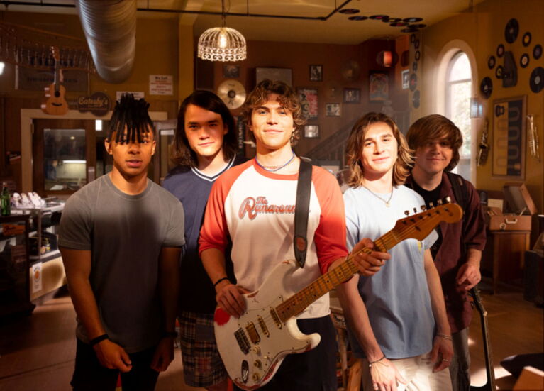 Ensemble cast stand in a music room with central figure holding an electric guitar