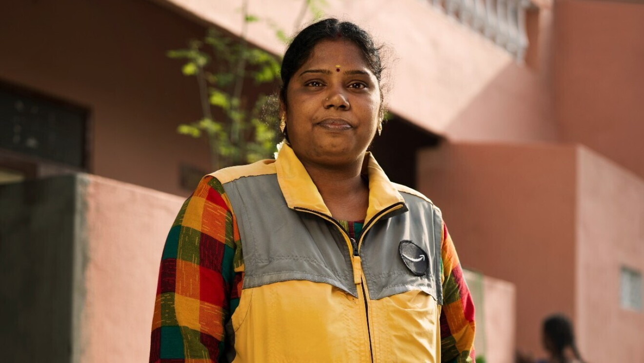 Keerthika  es una mujer india mirando a cámara. De fondo hay una casa de color rosa. La mujer va con una camisa estampada de cuadros de color rojo, amarillo y verde y lleva un chameco amarillo y gris con el logo de Amazon. 