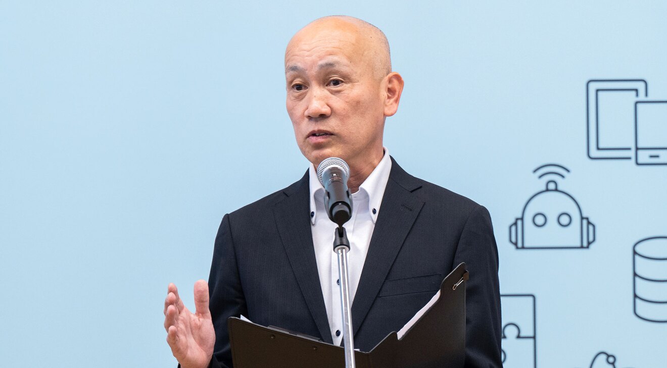 水色の背景の前に立ち、マイクを使用してスピーチをする人物。黒のスーツに白いボタンダウンシャツを着用し、左手で黒いクリップボードを開いている。背景には、タブレットやロボット、データベースなどを表す線画アイコンが描かれている。