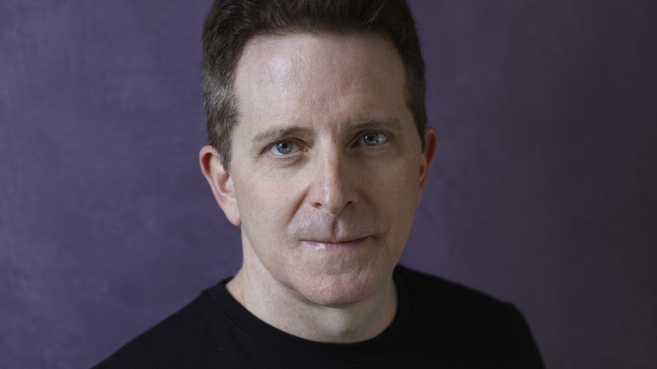 亚马逊作者兰德勒j .拿破仑盯着相机。他有黑色头发,蓝眼睛,穿着一件黑色的毛衣。背景是紫色。