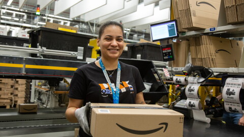 A young woman with brown hair and eyes is smiling, while holding a parcel in her hands working in a fulfillment center 